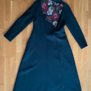 Unik & Stilfull Mörkgrön Lång Kappa med Broderade Blommor – Strl 38 - Gör ett riktigt statement i höst och vinter med denna exklusiva långkappa från Raslov! En dramatiskt vacker, mörkgrön kappa i figursmickrande modell med asymmetrisk omlottknäppning som ger ett modernt och sofistikerat intryck. Det som verkligen fångar ögat är det broderade blommönstret i röda, lila och gröna toner på framsidan – en unik och konstnärlig detalj som förvandlar kappan till ett riktigt blickfång. 🔹 Storlek: 38 🔹 Längd: ca 135 cm 🔹 Långa ärmar