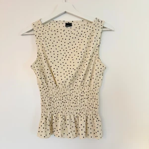 Prickig ärmlös topp från Gina Tricot - Supersöt topp från Gina Tricot i cremevit med svarta prickar. Toppen har smockad midja och volangkant nedtill som ger en smickrande siluett. Strl m men passar även s och xs i och med midjan🌸