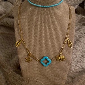 Två halsband med turkosa och guldiga detaljer - Ett guldigt kedjehalsband med berlocker som snäcka, sjöstjärna, elefant och en turkos fyrklöver. Perfekt för dig som gillar färg och unika detaljer. Medföljer även ett matchande armband i små turkosa pärlor 🩵