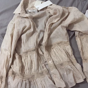 Beige blus med spets och broderi - Supersöt beige blus från By Malina med broderade detaljer och spetsinslag. Blusen har volang vid ärmslut och midja, knappar framtill och långärmad design. Perfekt för dig som gillar romantisk och bohemisk stil.