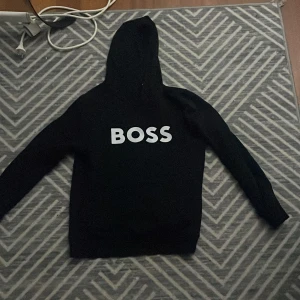 Svart hoodie från BOSS med tryck - Svart hoodie från BOSS i storlek S med stor vit BOSS-logga på bröstet. Tröjan har huva och långa ärmar, perfekt för chill dagar. Materialet känns mjukt och skönt mot huden. Enkel och stilren design som passar till jeans eller joggers. PRIS KAN DISKUTERAS
