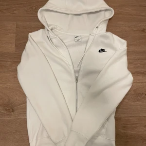 Vit hoodie med dragkedja från Nike - Snygg vit hoodie från Nike med svart logga på bröstet. Tröjan har huva med snörning, hel dragkedja och två fickor framtill. Perfekt för en chill och sportig stil. Mjuk och skön bomullsblandning.