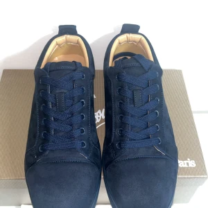 Mörkblå mocka sneakers från Louboutin - Snygga mörkblå sneakers i mocka från Louboutin med klassisk röd sula och broderad logga på hälen. Skorna har rund tå, platt sula och snörning. Insidan är fodrad i beige läder för extra komfort. Perfekta för dig som vill sticka ut med exklusiv stil.