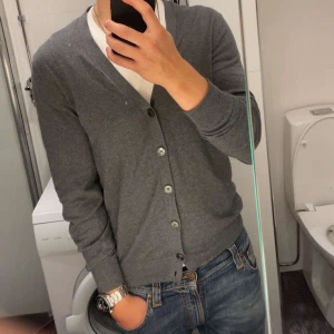 Grå kofta från Massimo Dutti - Säljer en elegant grå kofta från Massimo Dutti, tillverkad i en lyxig blandning av bomull, silke och kashmir. Koftan har knappar framtill och ribbade muddar vid ärmslut och nederkant. Perfekt för en stilren look.