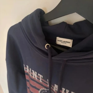 Saint Laurent marinblå hoodie med tryck - Säljer en marinblå hoodie från Saint Laurent med stort vintageinspirerat tryck i vitt och rött framtill. Tröjan har huva med snörning och ribbade muddar. Perfekt för dig som gillar streetstyle och exklusiva märken.