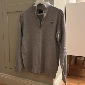 Grå half zip tröja Lyle & Scott - Grå långärmad tröja från Lyle & Scott Junior med half zip och ståkrage. Tröjan har ett litet broderat gult logomärke på bröstet och är tillverkad i mjuk bomull. Perfekt för chill dagar och snygg till jeans eller joggers. OBS: liten röd fläck på ryggen, syns inte vid andvändning. 