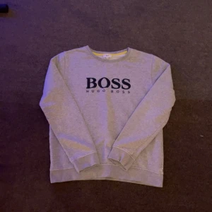 Grå sweatshirt från Hugo Boss - Snygg grå sweatshirt från Hugo Boss med svart logga framtill. Tröjan har rund halsringning och långa ärmar. Tillverkad i mjuk bomullsmix som är skön att ha på sig. Perfekt för dig som gillar stilrena och klassiska plagg med en modern touch.