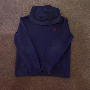 Mörkblå hoodie från Polo Ralph Lauren - Säljer en mörkblå hoodie från Polo Ralph Lauren i storlek M-L. Tröjan har klassisk huva, långa ärmar och en röd broderad logga på bröstet. Perfekt för dig som gillar stilren och enkel design med en touch av lyx.