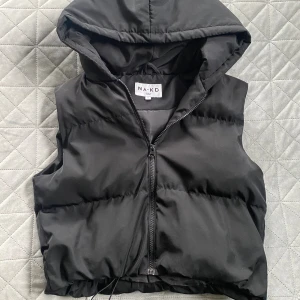 Dunväst med huva från NA-KD - Svart dunväst från NA-KD med huva och dragkedja framtill. Västen har en quiltad look och dragsko nertill för justerbar passform. Kort modell. Som ny!