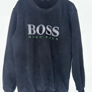  Hugo Boss sweatshirt    - Svart sweatshirt från Hugo Boss med stor BOSS-logga i vitt och grönt framtill. Tröjan har rund halsringning, långa ärmar och är tillverkad i mjuk bomullsmix. Perfekt för en chill och stilren look.