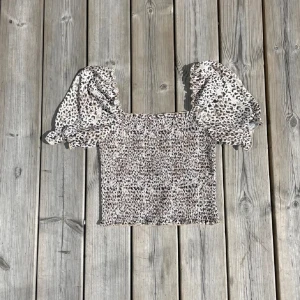 Leopardmönstrad blus från NA-KD - Säljer en söt blus från NA-KD med puffiga korta ärmar och leopardmönster i beige, svart och grå toner. Toppen är smockad över hela kroppen och har volangdetaljer vid ärmslut och nederkant. Perfekt för dig som gillar trendiga och djuriska prints!