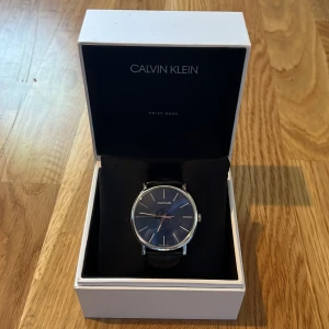 Calvin Klein klocka - Stilren klocka från Calvin Klein med rund urtavla i mörkblått och silverdetaljer. Svart läderarmband med spänne där Calvin Klein är ingraverat. Kommer i original ask. Perfekt för dig som gillar minimalistisk design och snygga accessoarer. Skicket är bra, endast lite repor på glaset. Nypris låg på typ 2200 om jag minns rätt men är inte helt säker då jag inte hittar kvittot. Batteri behövs bytas. 