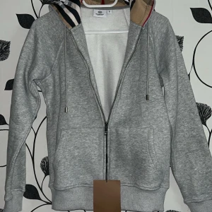 Grå hoodie från Burberry med dragkedja - Säljer en grå hoodie från Burberry med klassisk dragkedja och snörning i huvan. Insidan av huvan har Burberrys ikoniska randiga mönster i beige, svart och vit. Tröjan har två fickor framtill och ribbade muddar vid ärmslut och nederkant. Perfekt för chill dagar.