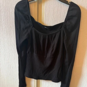 Svart ribbad långärmad blus - Säljer en svart ribbad blus från Gina Tricot med lång ärm och snygg fyrkantig ringning. Toppen har en lätt rynkad detalj vid bysten och är croppad i modellen. Perfekt för dig som gillar stilrena och trendiga plagg.