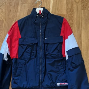 Tommy Hilfiger Jacka - Snygg vindjacka från Tommy Jeans i marinblått, rött och vitt. Jackan har dragkedja, stora fickor framtill och tydlig logga på ryggen. Tillverkad i polyester med meshfoder för extra komfort. Perfekt för dig som gillar sportig och färgstark stil. Den är aldrig använd och i perfekt skick. Sitter som en xs i storleken.