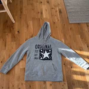 Grå hoodie med stjärntryck - Säljer en grå hoodie med huva och snörning, tryck med stjärnor och texten 'Original Fit Est. 2024' på framsidan. Tröjan har en stor magficka och långa ärmar. Perfekt för chill dagar och enkel att matcha med jeans eller mjukis.