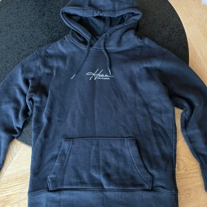 Svart hoodie från Hollister - Svart hoodie från Hollister med vit logga på bröstet. Tröjan har huva med snörning, stor magficka och ribbade muddar vid ärmslut och nederkant. Perfekt för chill dagar och har en klassisk, loose passform.