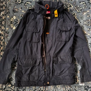 Vindjacka från Parajumpers - Svart vindjacka från Parajumpers med flera stora fickor framtill, huva och snygga detaljer som gul rem vid kragen. Jackan har meshfoder för ventilation och stängs med både dragkedja och knappar. Perfekt för dig som gillar funktion och stil.