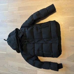 Svart pufferjacka från Canada Goose - Svart Mc maillan jacka från Canada Goose med huva och snyggt ton-i-ton märke på ärmen. Jackan har quiltad design, dragkedja framtill och ribbade muddar vid ärmslut. Perfekt för kalla dagar och har en clean, modern look.