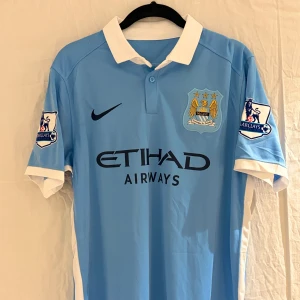 Manchester City De Bruyne tröja 2015/2016 - Manchester City retro från säsongen 2015/2016 med De Bruyne nummer 17 på ryggen. Tröjan är ljusblå med vita detaljer, korta ärmar och klassisk krage. Nike-logga, Premier League-patch på båda ärmar och broderat klubbmärke på bröstet. Materialet är lätt och ventilerande polyester.