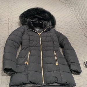 Svart pufferjacka med fuskpäls från Lindex - Svart pufferjacka från Lindex med fluffig fuskpäls på huvan och guldfärgade dragkedjor. Jackan har quiltad design, två fickor med dragkedja och lång ärm. Perfekt för kalla dagar med sin varma och mysiga känsla. Storlek S passar M