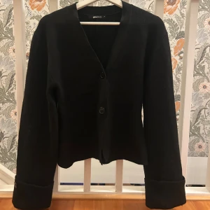 Svart kofta från Gina Tricot - Stilren svart kofta från Gina Tricot med v-ringning och två knappar framtill. Koftan har långa ärmar med breda muddar och en rak, något kortare passform. Den är helt ny endast provad, beställde hem i fel storlek. Tröjan är i storlek M men passar även S🩷