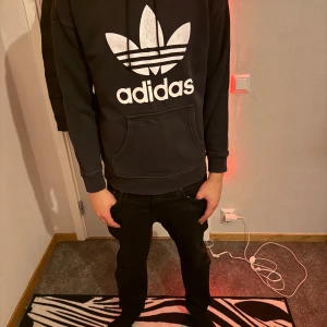 Svart Adidas hoodie med stor logga - Säljer en svart hoodie från Adidas med stor vit trefoil-logga och text på bröstet. Tröjan har huva med snörning, känguruficka och ribbade muddar. Perfekt för dig som gillar streetwear och sportig stil.