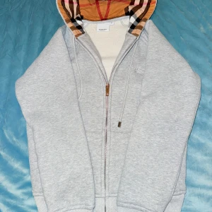 Grå Burberry hoodie med rutig huva - Säljer en grå hoodie från Burberry med dragkedja och klassisk rutig huva i beige, svart och röd. Tröjan har långa ärmar, ribbade muddar och snörning vid huvan. Perfekt för dig som vill ha en snygg och bekväm streetstyle-look. Pris går att diskutera