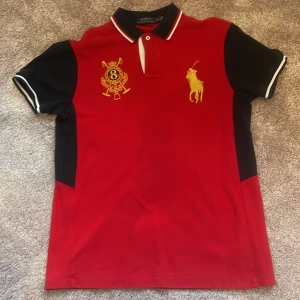Röd pikétröja Polo Ralph Lauren - Snygg röd pikétröja från Polo Ralph Lauren med svarta detaljer på ärmar och sidor. Tröjan har broderade emblem och en stor gul Polo-logga på bröstet samt siffran 8 på ryggen. Kragen har vita och svarta ränder och modellen är custom fit.