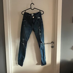 Blå skinny jeans från Lager 157 - Säljer ett par blå skinny jeans från Lager 157 med slitna detaljer och hål på benen. Jeansen har klassisk femficksdesign, knappstängning och är tillverkade i stretchigt denim för skön passform. 