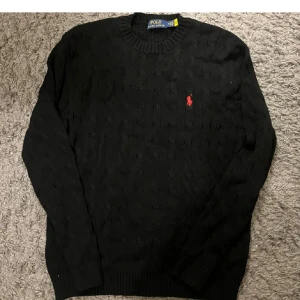 Svart stickad tröja från Polo Ralph Lauren - Svart stickad tröja från Polo Ralph Lauren med klassiskt kabelstickat mönster och röd broderad logga på bröstet. Rund hals och långa ärmar med ribbade muddar. Perfekt för dig som gillar stilrena och tidlösa plagg.