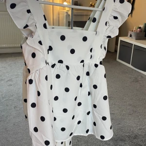 Vit prickig kortklänning med volang - Supersöt vit topp med svarta prickar och volangdetaljer på axelbanden. Klänningen har en knytning i ryggen. Säljer då den aldrig kommit till användning.