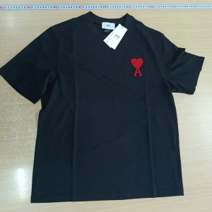 Svart t-shirt från Ami med rött hjärta - Svart t-shirt från Ami Alexandre Mattiussi med klassisk passform och rund hals. På bröstet finns ett rött broderat hjärta med bokstaven A. T-shirten är i mjuk bomull och har korta ärmar. Perfekt för en clean och stilren look.