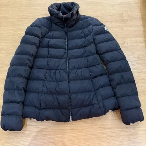 Svart Moncler pufferjacka Äkta - Svart pufferjacka från Moncler med hög krage och klassisk logga på ärmen. Jackan har quiltad design, dragkedja framtill och är tillverkad i polyester med dunfyllning. Perfekt för kalla dagar och ger en clean, stilren look. Pris kan diskuteras vid snabbaffär, 100% äkta!