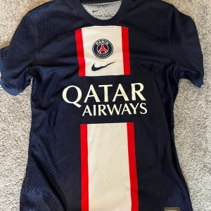 PSG matchtröja Nike blå röd vit - Paris Saint-Germain fotbollströja från Nike med klassisk design. Marinblå bas med bred vit vertikal rand och röda kanter på framsidan, PSG-logga och Qatar Airways-tryck. Kortärmad, rund hals och tillverkad i lätt, ventilerande Dri-FIT-material.
