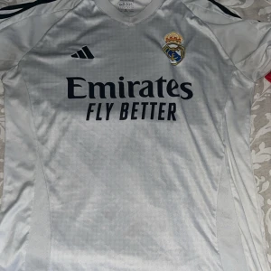 Real Madrid Vini Jr. 7 fotbollströja - Säljer en vit Real Madrid fotbollströja från Adidas med Vini Jr. och nummer 7 på ryggen. Tröjan har korta ärmar, klubbmärke på bröstet och sponsortryck 'Emirates Fly Better' framtill. Materialet är lätt och andas, perfekt för match eller träning. Kan diskutera om priset!