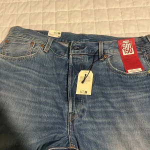 Levi's 501 blå jeans med slitningar - Säljer ett par klassiska Levi's 501 jeans i ljusblå tvätt med snygga slitningar och hål på benen. Modellen har raka ben, knappgylf och fem fickor. Perfekt för dig som gillar en avslappnad och cool stil.