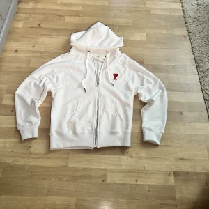 Vit hoodie från Ami med rött hjärta - Vit hoodie från Ami med dragkedja, huva och snörning. På bröstet finns ett broderat rött hjärta med bokstaven A. Tröjan har ribbade muddar och fickor framtill. Materialet är mjuk bomull och logga broderad på ärmen.