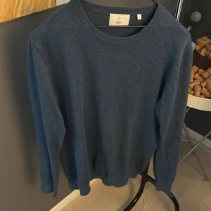 Mörkblå stickad tröja från Bläck - En enkel och stilren mörkblå stickad tröja från Bläck i storlek M. Tröjan har rund hals, långa ärmar och ribbade muddar vid hals, ärmslut och nederkant. Perfekt för dig som gillar klassisk och clean look.