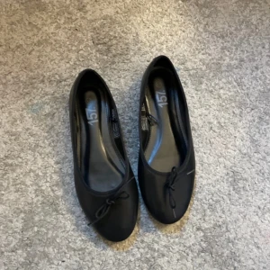 Svarta ballerinaskor från 157, strl 37 - Klassiska svarta ballerinaskor från 157 i storlek 37. Skorna har rund tå, platt sula och en liten rosett framtill. Perfekta för en clean och enkel look. Tillverkade i syntetmaterial med mjuk innersula för extra komfort.