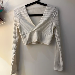 Vit croppad omlottopp  - Säljer en vit croppad omlottopp från SHEIN i ribbad bomullsmix. Toppen har lång ärm, v-ringad omlottdesign och sitter snyggt med sin slim passform. Perfekt för dig som gillar stilrena och trendiga plagg med lite edge.