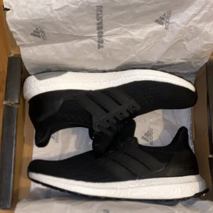 Adidas Ultraboost svarta sneakers 42 - Svarta Adidas Ultraboost sneakers med stickad ovandel och klassiska tre ränder på sidan. Skorna har en vit, responsiv Boost-sula och svart yttersula. Snörning framtill och rund tå. Perfekta för dig som gillar stilren och sportig look.