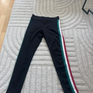 Svarta leggings med färgglada ränder - Säljer ett par svarta leggings med bred resår i midjan och snygga ränder i grönt, vitt och rött längs sidorna. Perfekta för dig som gillar sportig stil och vill ha något bekvämt och trendigt. Materialet är mjukt och stretchigt för maximal komfort.
