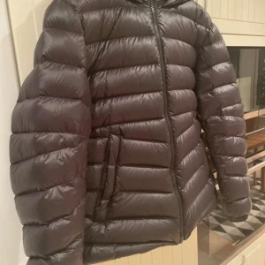 Svart Colmar pufferjacka med huva - Säljer en svart pufferjacka från Colmar Originals med huva och dragkedja framtill. Jackan har ett stilrent, quiltat mönster och logotyp på ärmen. Perfekt för kalla dagar och har en snygg, modern look som passar till streetwear. Men den är liten i storleken