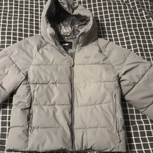 Grå pufferjacka från Pull&Bear - Snygg grå pufferjacka från Pull&Bear med huva och dragkedja framtill. Jackan har två fickor med dragkedja och quiltad design. Perfekt för kalla dagar och har en modern, boxig passform.