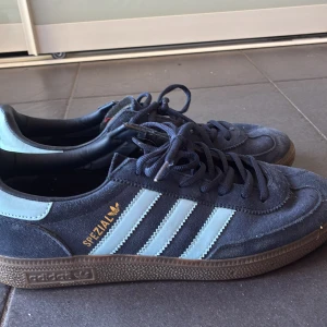Adidas Spezial sneakers i mörkblå mocka - Säljer ett par Adidas Spezial sneakers i mörkblå mocka med ljusblå ränder och detaljer. Skorna har klassisk gummisula, snörning och rund tå. Perfekta för dig som gillar retrostil och streetwear. Ikonisk Spezial-logga i guld på sidan.