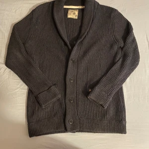 Mörkgrå ribbad kofta Jack & Jones - Säljer en mörkgrå ribbad kofta från Jack & Jones Vintage Cloth i storlek medium. Koftan har två framfickor, knappar hela vägen och en bred krage. Tillverkad i bomull med grovstickad känsla, perfekt för lager på lager under kyliga dagar.