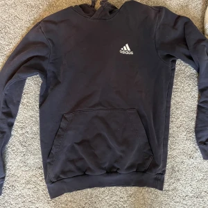 Svart hoodie från Adidas - Svart hoodie från Adidas med klassisk logga i vitt på bröstet. Tröjan har huva med snörning, stor magficka och långa ärmar. Perfekt för chill eller träning, enkel och stilren design i mjuk bomull.