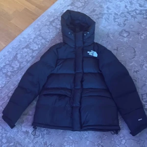 Svart dunjacka från The North Face M - Säljer en svart dunjacka från The North Face i storlek M. Jackan har huva, dragkedja och snörning nedtill. Klassisk pufferstil med logga på bröstet och ryggen. Perfekt för kalla dagar och riktigt snygg streetstil.