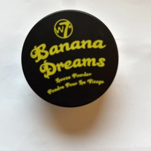 W7 Banana Dreams Loose Powder - Banana Dreams Loose Powder från W7 är ett ansiktspuder i en rund svart burk med gul text. Pudret har en ljusgul ton som ger en matt finish och hjälper till att ljusa upp och fixera din makeup. Perfekt för att få en fräsch och jämn look. Endast testad